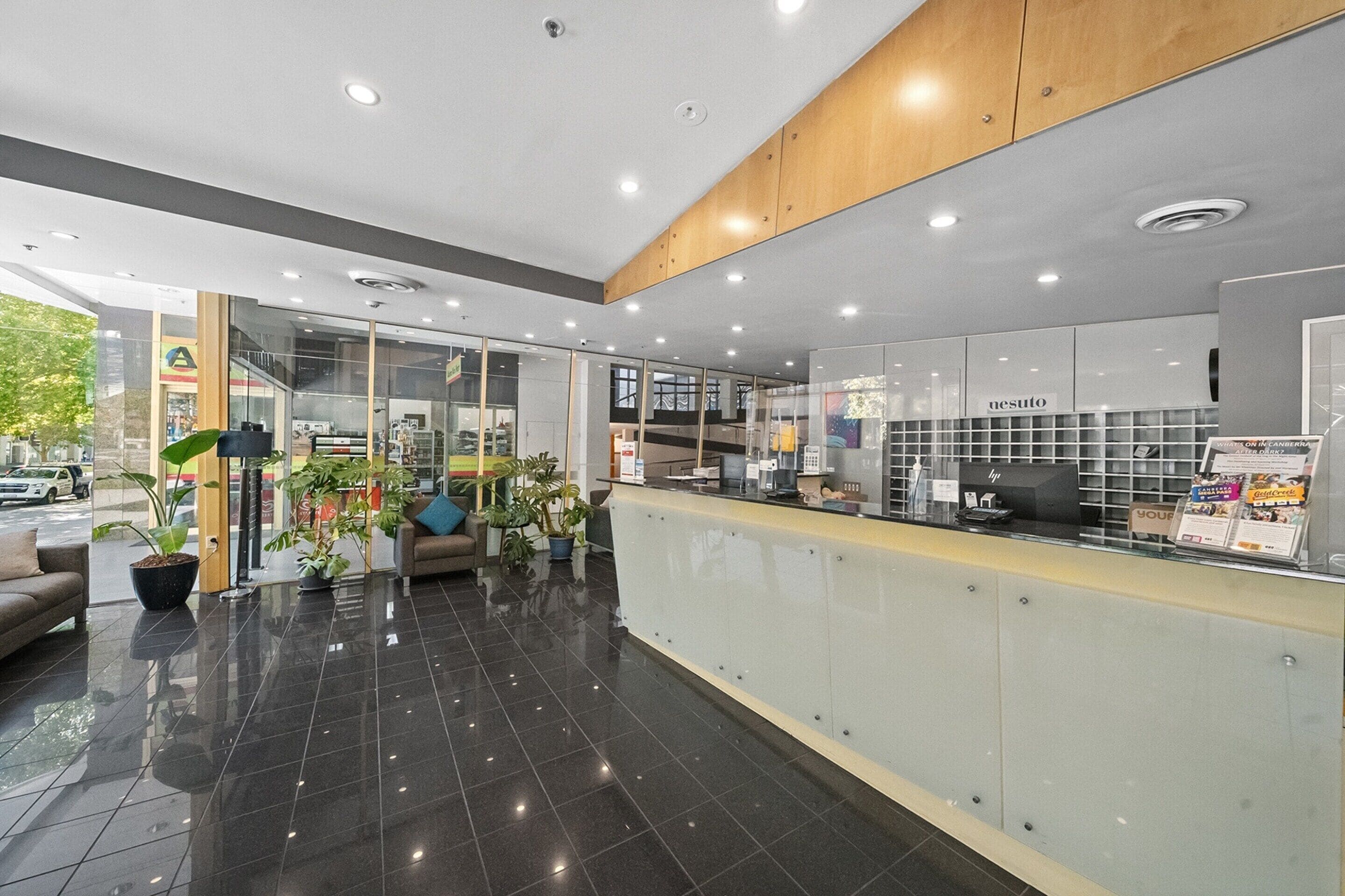A modern reception area with a glass partition, a reception desk, and a seating area with a sofa and potted plants.