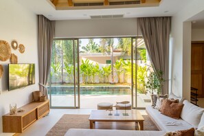 Ein modernes Wohnzimmer mit einer großen Schiebeglastür, die zu einem Poolbereich führt, eine Couch mit Kissen, ein Couchtisch und dekorative Wandbilder.