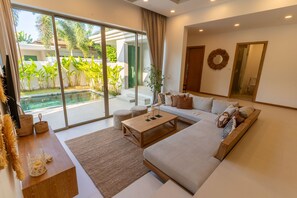 A modern living room with a large glass door leading to a pool, a sofa with cushions, a coffee table, and a small side table with decorative items. - Private Pool Villa in the Heart of Bang Tao (ต.เทพกระษัตรีย์)