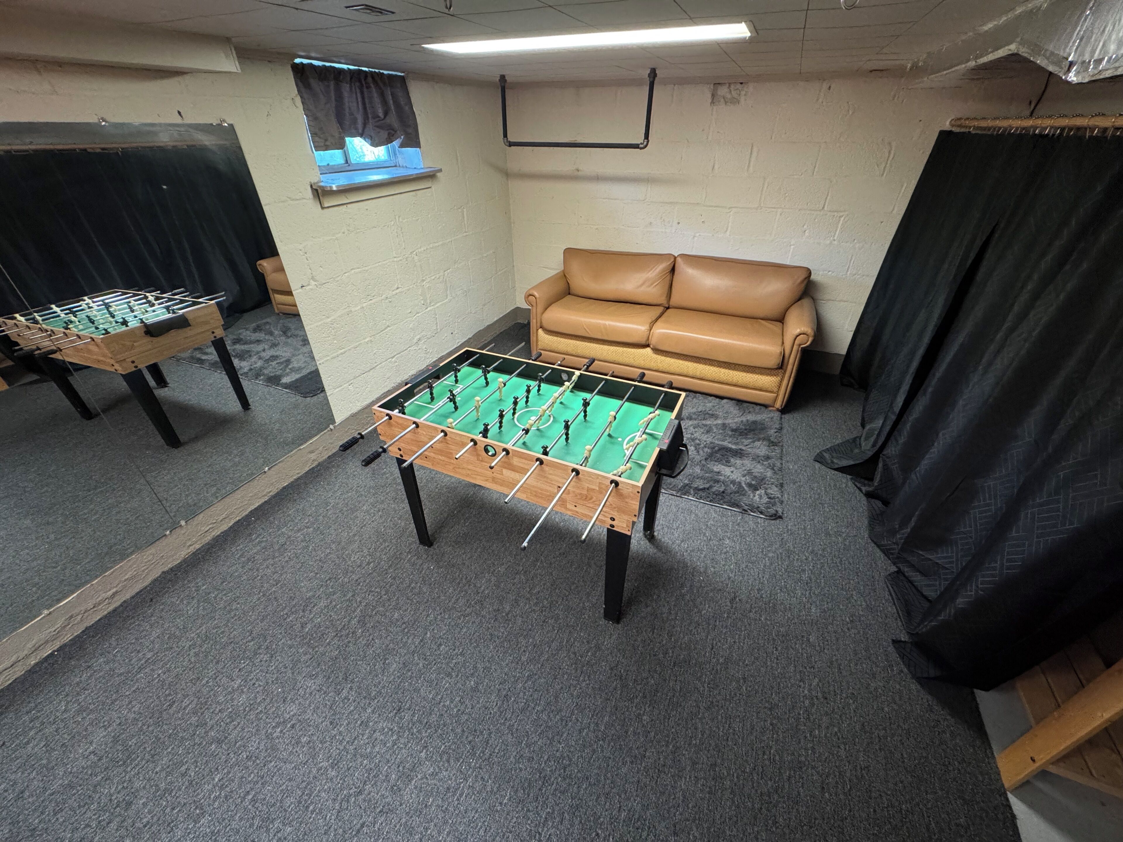 Una habitación con dos mesas de futbolín, un sofá de cuero marrón y un ventanal con cortinas.