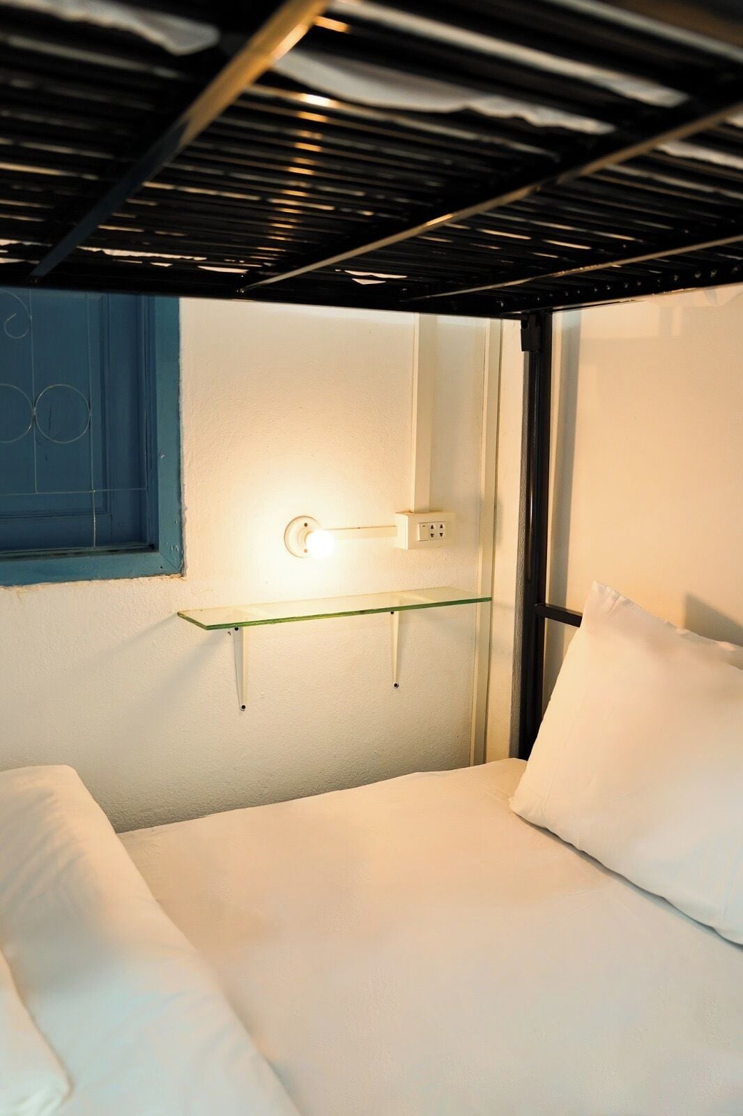 Un lit superposé avec une étagère au-dessus, une prise de courant au mur et une fenêtre avec un cadre bleu.
