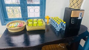 A table with a coffee maker, a basket of snacks, a tray of green cups, and a stack of bottled water. - Keokhounmuang Hostel (Luang Prabang)