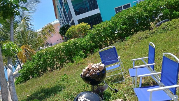 A grassy outdoor area with a grill, chairs, and a pool in the background.