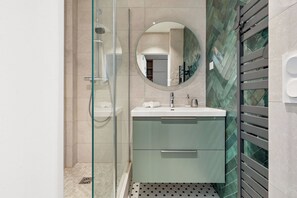 A modern bathroom with a glass shower enclosure, a round mirror, and a sink with a green cabinet. - Chic Flat near Opéra & Marais (Paris)