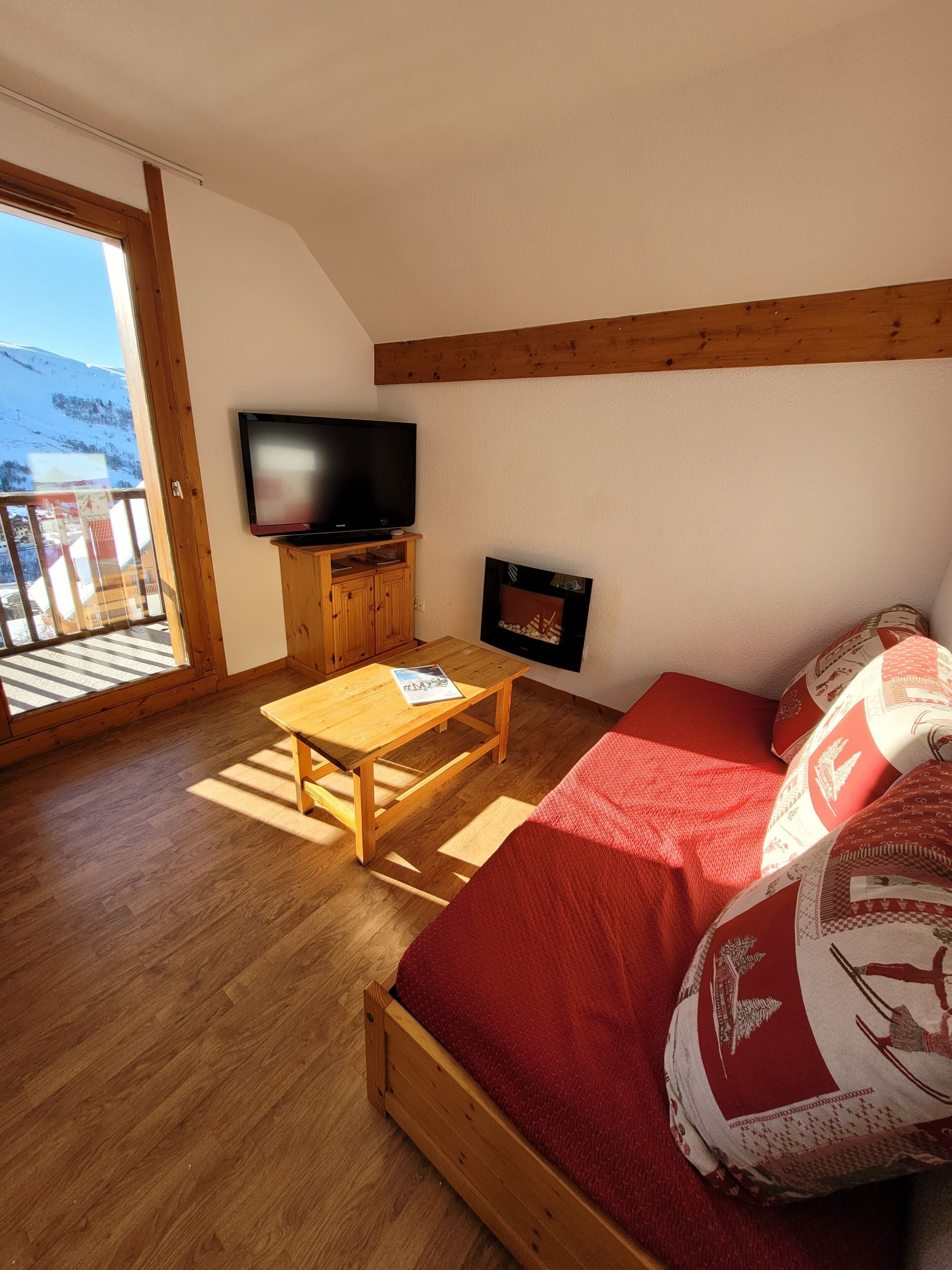 Ein Zimmer mit Holzboden, einem Sofa mit roten Kissen, einem an der Wand montierten Fernseher und einem Kamin.