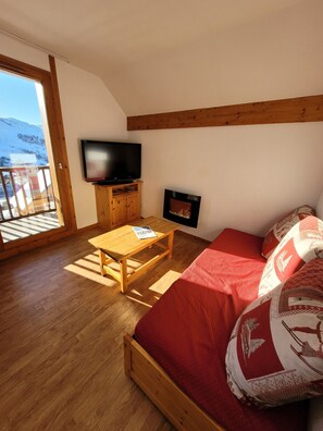 Ein Zimmer mit Holzboden, einem Sofa mit roten Kissen, einem an der Wand montierten Fernseher und einem Kamin.