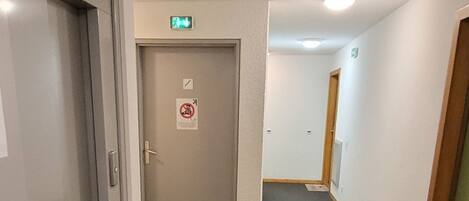 Un pasillo con piso alfombrado, una puerta con letrero de no fumar y un detector de incendios en el techo.