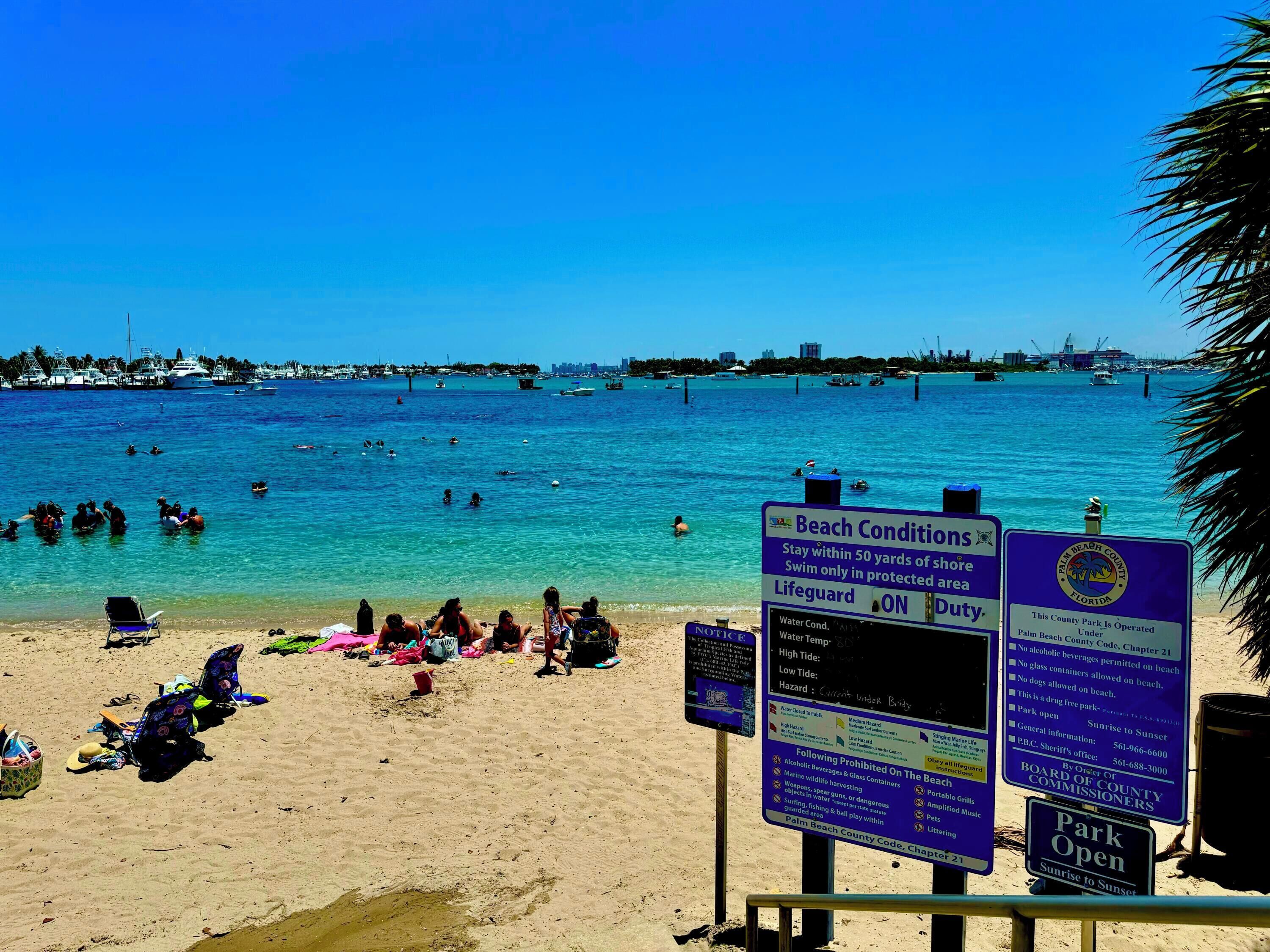 A beach with clear blue water, people swimming and sunbathing, and multiple signs with beach conditions and regulations.