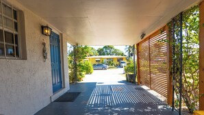 A covered porch with a blue door, a yellow car parked in the background, and a wooden fence on the right. - Rent The Crafty Mermaid, 2-bedroom house, yard, near Blue Heron dive area (West Palm Beach)
