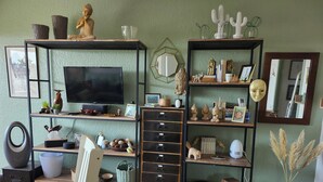 Un salon comprenant un téléviseur, des étagères ornées d’objets décoratifs, un fauteuil et un vase contenant des plantes séchées.