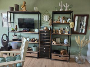 Ein Wohnzimmer mit Fernseher, Regalen mit Dekoration, einem Sessel und einer Vase mit Trockenpflanzen.