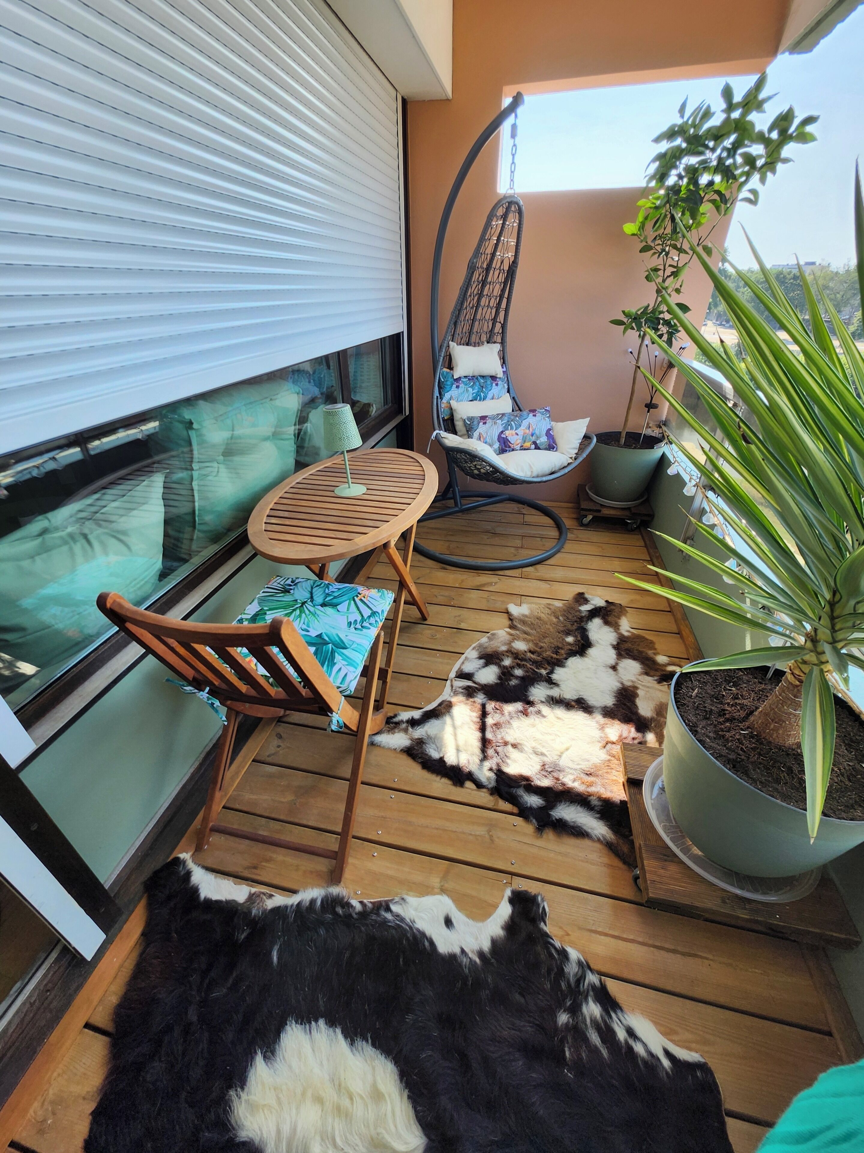 Un balcon avec une table en bois, un fauteuil suspendu, une plante en pot et un tapis en fourrure.
