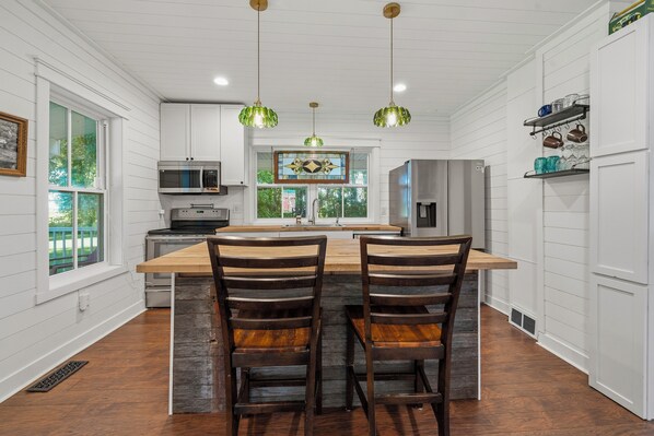 A kitchen with a central island, two barstools, hanging pendant lights, and a view of the outdoors through a window.