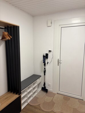 A room with a bench, a door, a white wall, a shelf with hanging items, and a vacuum cleaner. - Le Cosy - Apartment with swimming pool near Lake Gerardmer (Gérardmer)