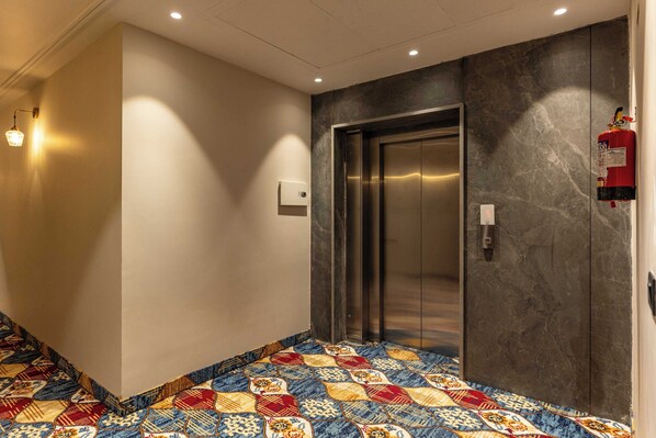 A hallway with a patterned carpet, an elevator, a fire extinguisher, and a wall-mounted light fixture. - Hotel City Klyde Inn (Amritsar)
