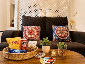 A living room with a dark sofa, two decorative pillows, a basket with snacks, a plant, and a candle.