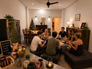 A group of people seated around a table in a well-lit room with a ceiling fan, a Christmas tree, and various snacks and drinks on the table.
