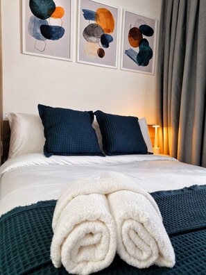 A neatly made bed with white linens, two dark blue pillows, and a rolled white towel on a dark green textured blanket. - Studio Brooklin, EXPO (São Paulo)