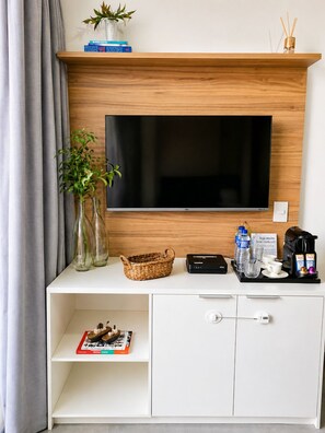 A modern hotel room with a flat-screen TV mounted on a wooden wall, a white console table with a basket, a coffee maker, and a small selection of books. - Studio Brooklin, EXPO (São Paulo)