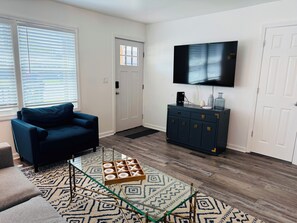 A living room with a dark blue sofa, a glass coffee table with a geometric patterned rug, a dark wooden cabinet with a flat-screen TV above it, and a white door in the background. - Chic, coastal, pet-friendly retreat located in Lower Township, NJ. (Lower Township)