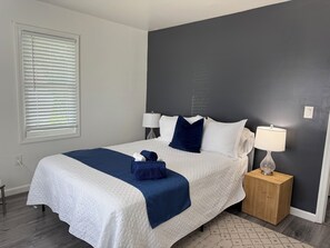 A neatly made bed with a white and blue quilt, two bedside lamps, a wooden nightstand with a lamp, and a window with blinds. - Chic, coastal, pet-friendly retreat located in Lower Township, NJ. (Lower Township)