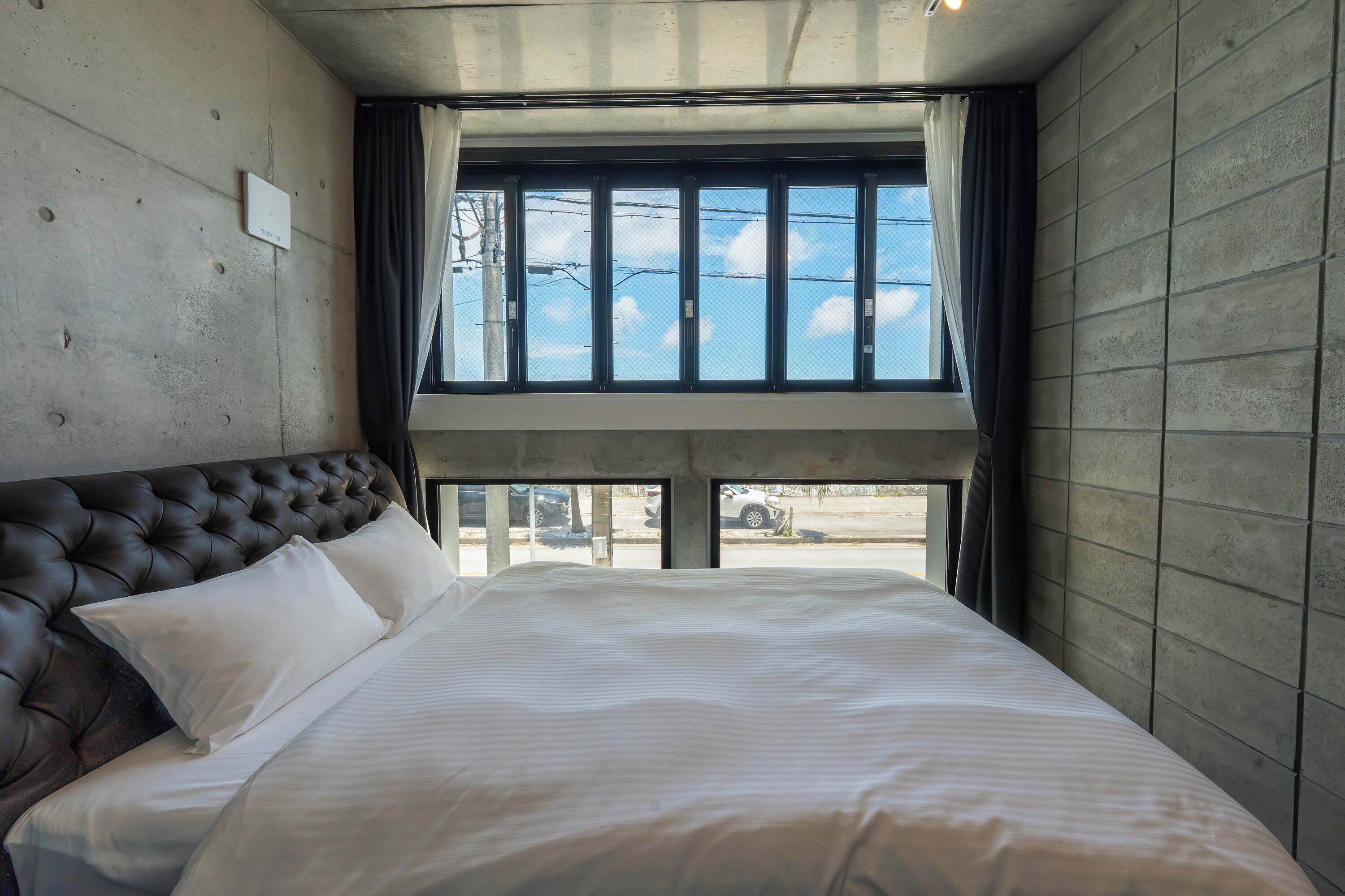 Quarto com uma cama grande, cabeceira e janela com vista para o céu e linhas de eletricidade.