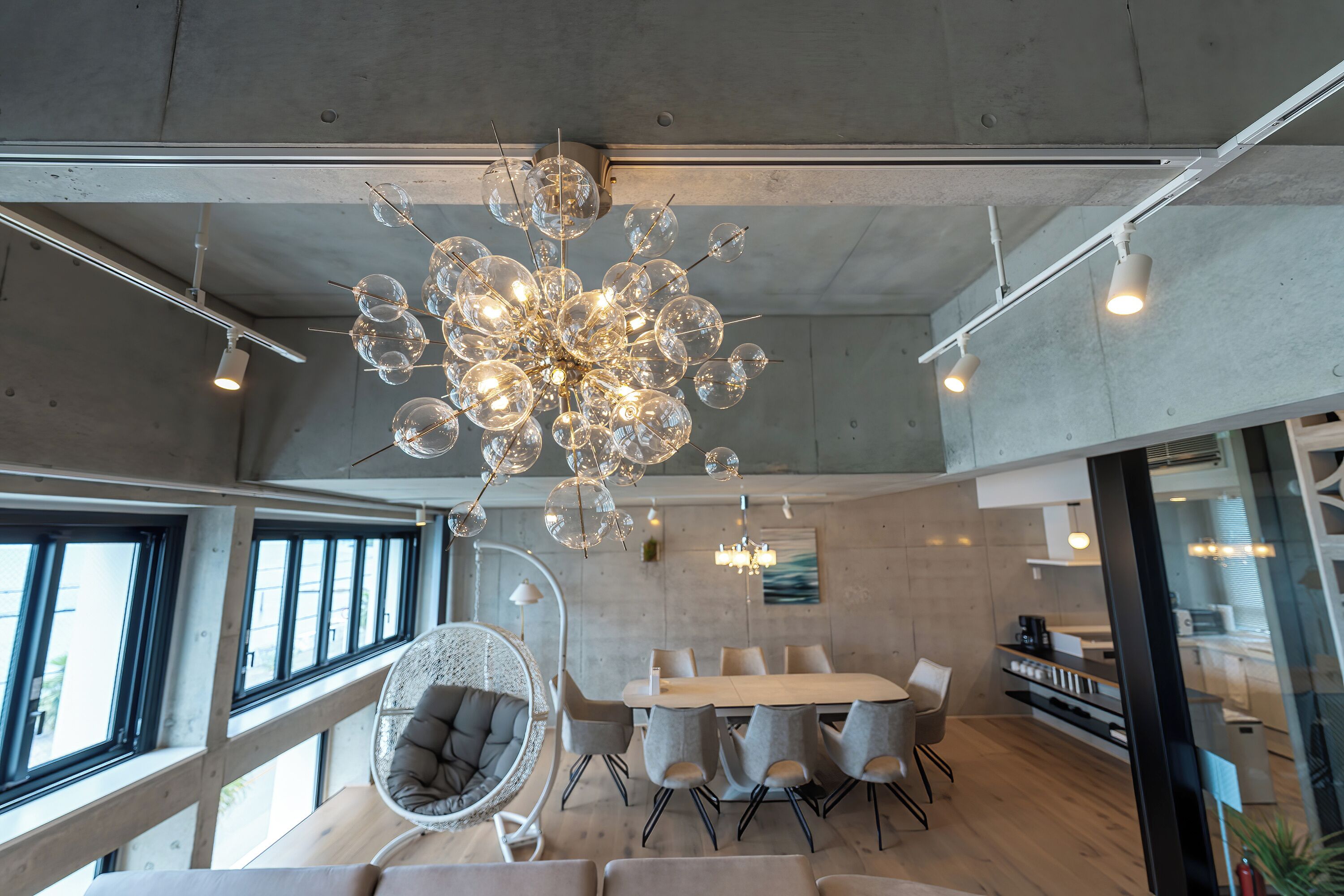 Um interior moderno com um lustre esférico único, uma área de refeições com mesa e cadeiras redondas, e uma cadeira de vime com almofada.