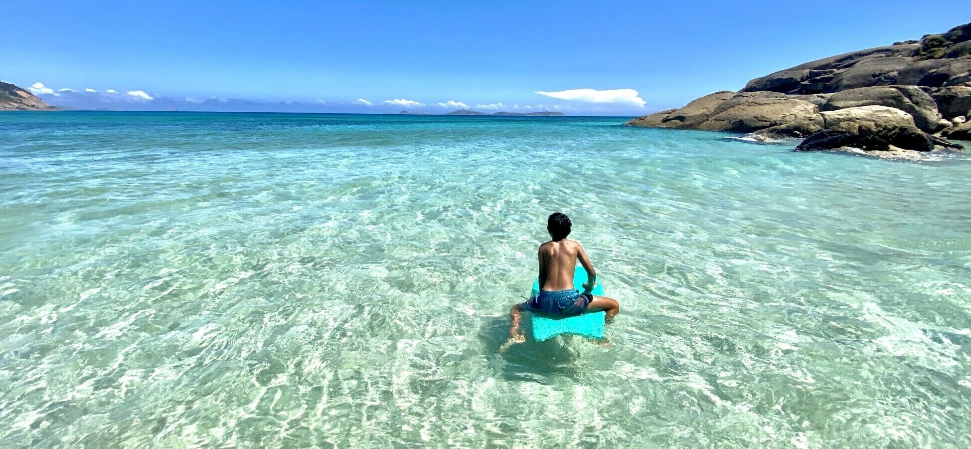 Una persona sentada en un flotador azul en agua turquesa cristalina, con una costa rocosa y montañas distantes.