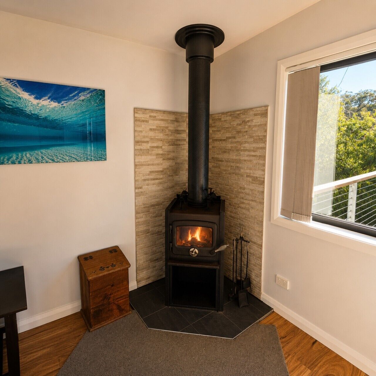 A room with a wood-burning stove, a window with curtains, a wooden chest, and a painting on the wall.