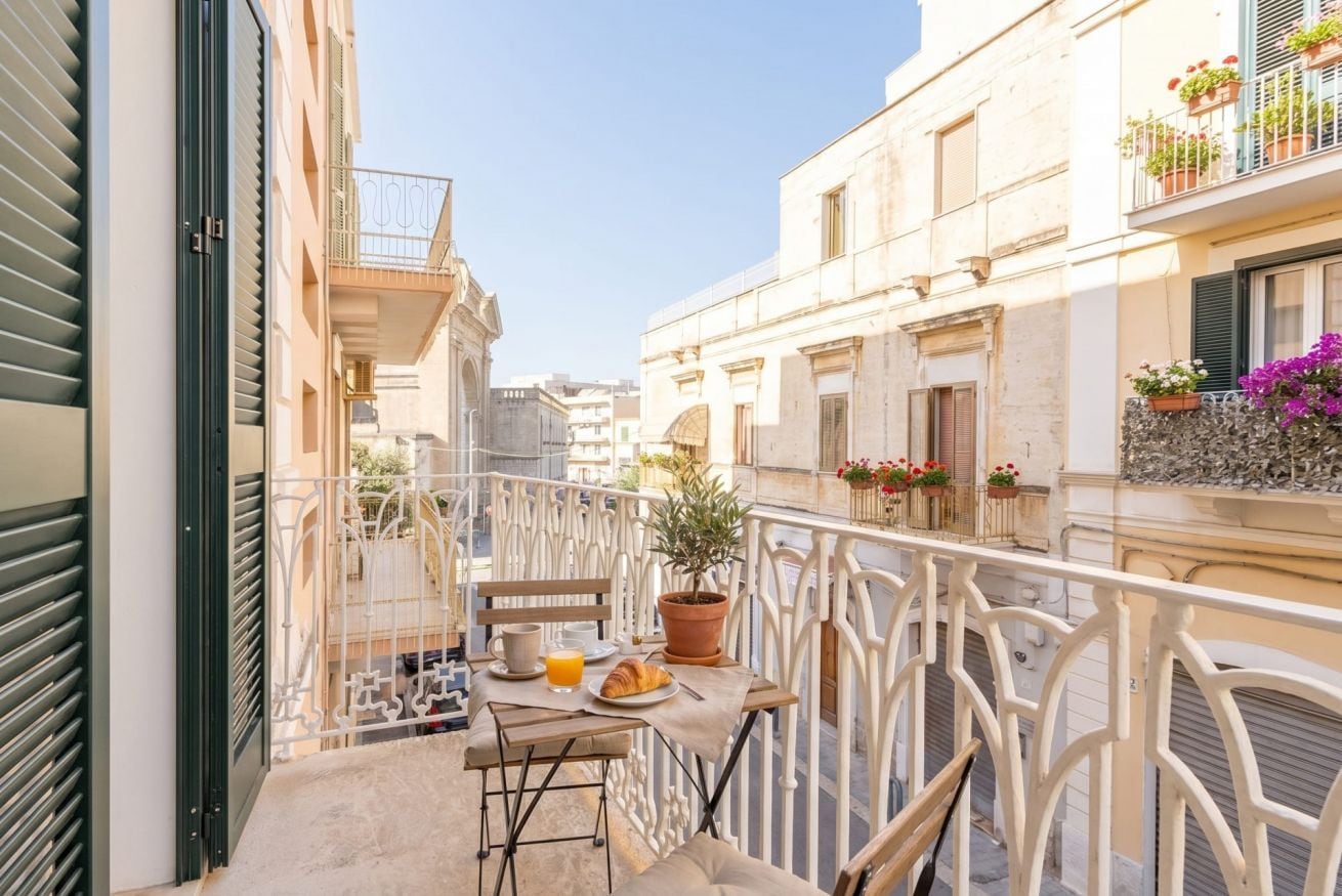 A balcony with a table set for breakfast, a potted plant, and a view of the street.