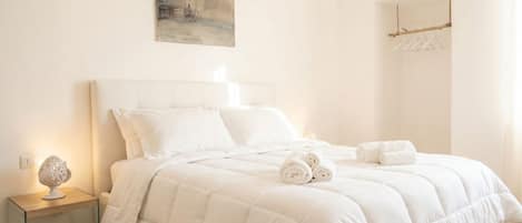 A neatly made bed with white bedding and rolled towels, a bedside lamp, and a painting on the wall.
