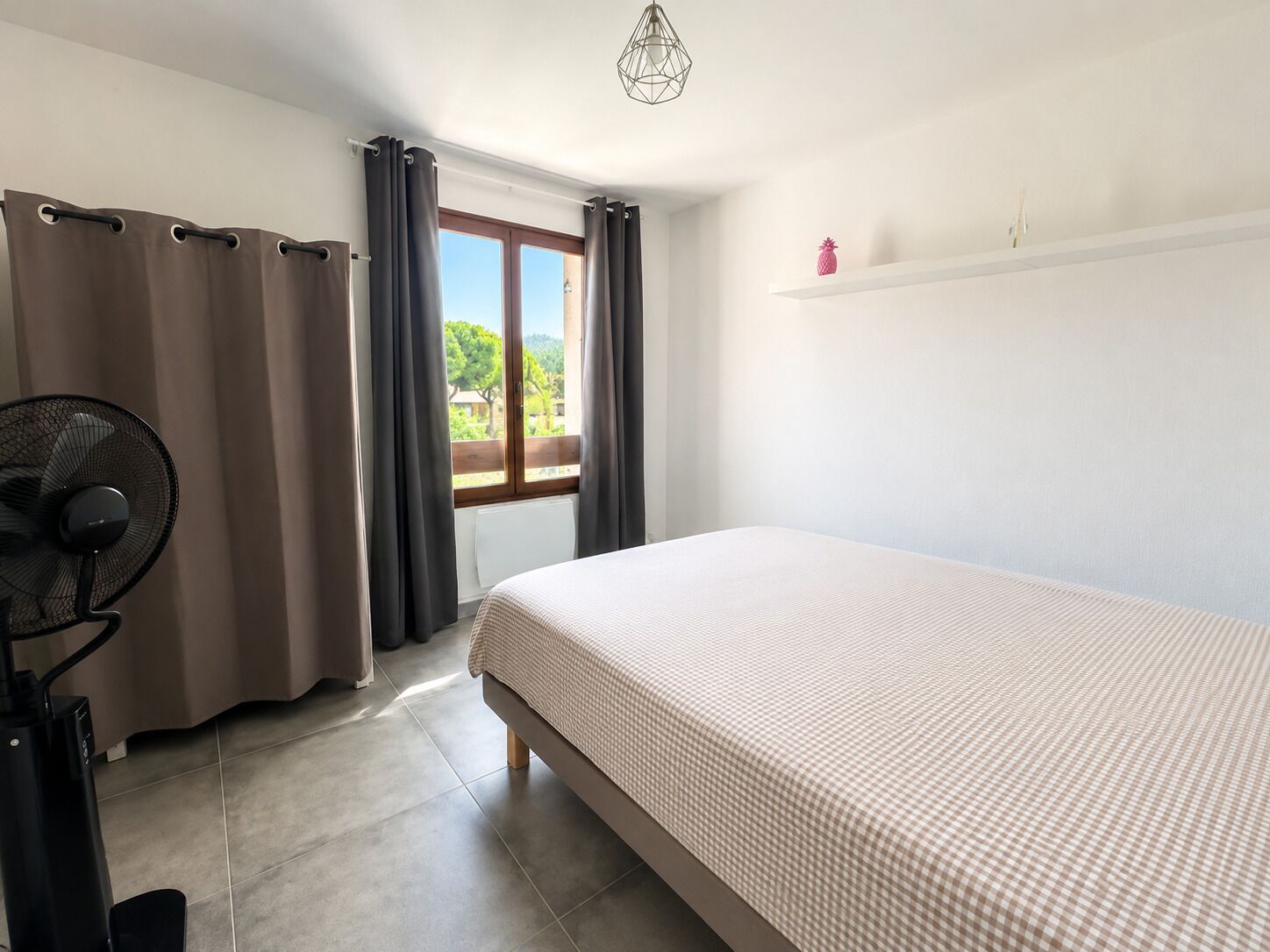 A bedroom with a bed, a standing fan, and a window with curtains.