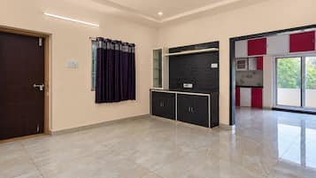A spacious room with a dark wooden door, a window with purple curtains, a black cabinet with a textured wall above, and a red and white cabinet with a glass door.