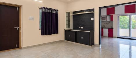 A spacious room with a dark wooden door, a window with purple curtains, a black cabinet with a textured wall above, and a red and white cabinet with a glass door.