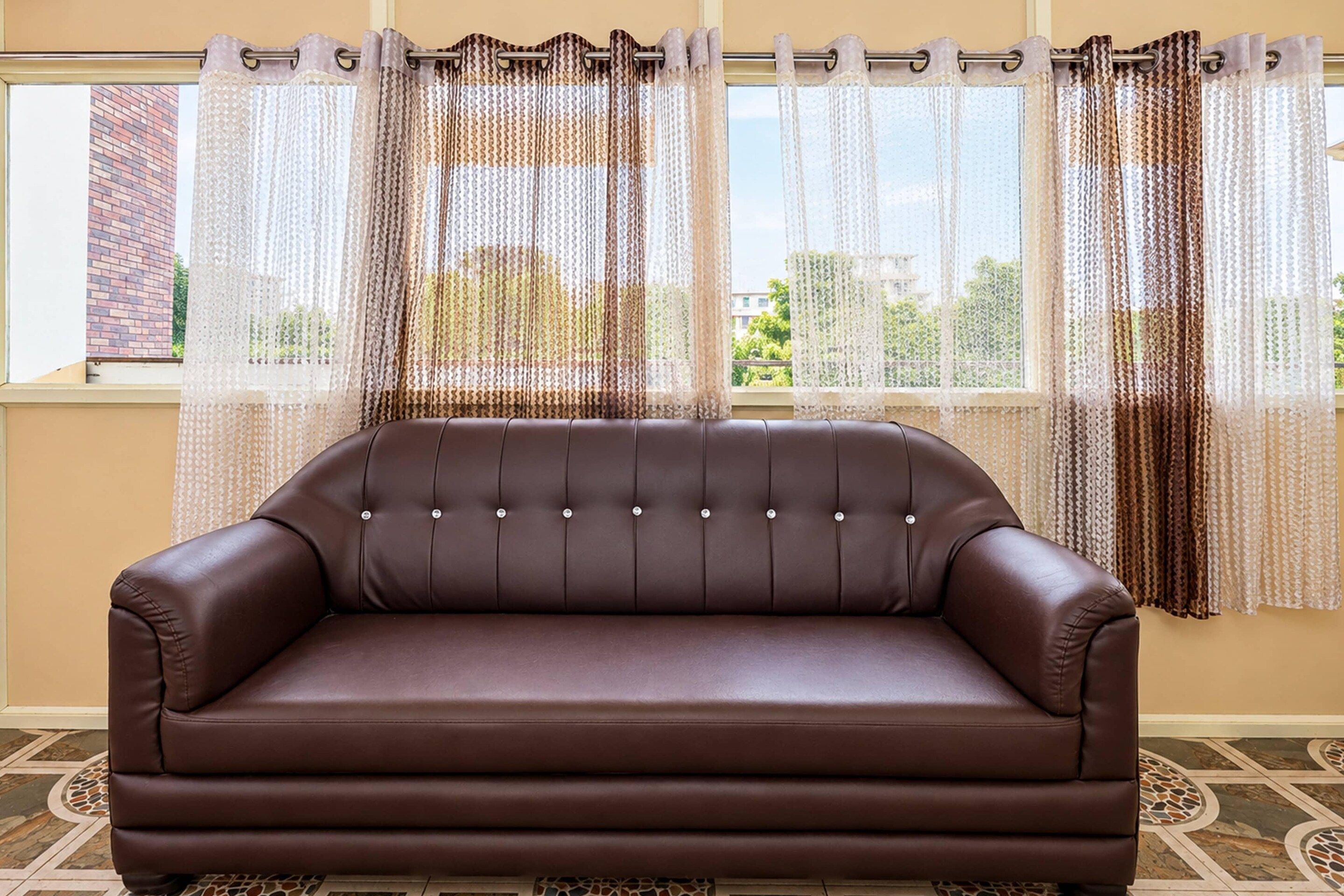 A brown leather sofa in a room with a patterned rug, a window with a view of buildings and greenery, and a curtain with a brown and white pattern.