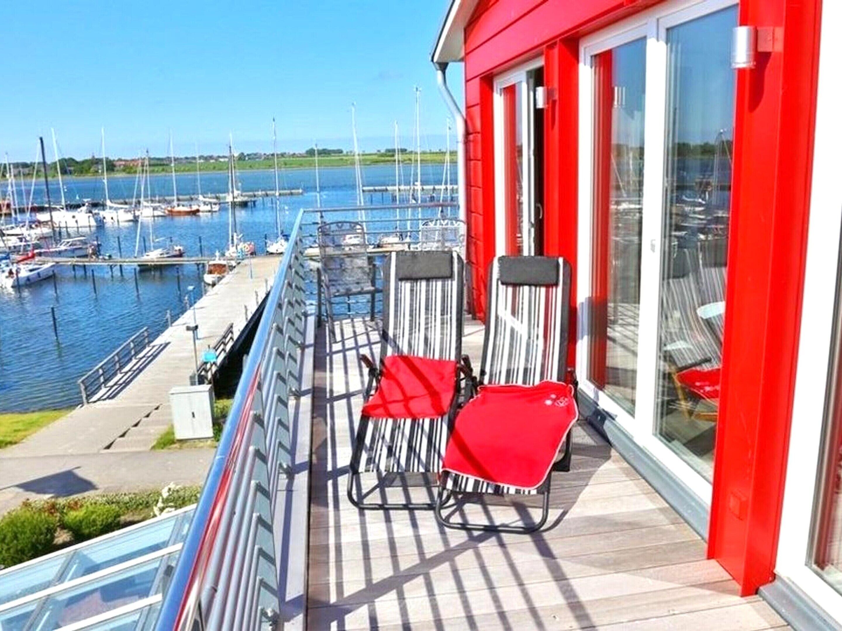 Ein Balkon mit zwei Stühlen, Blick auf einen Yachthafen mit Booten und ein rotes Gebäude mit großen Fenstern.