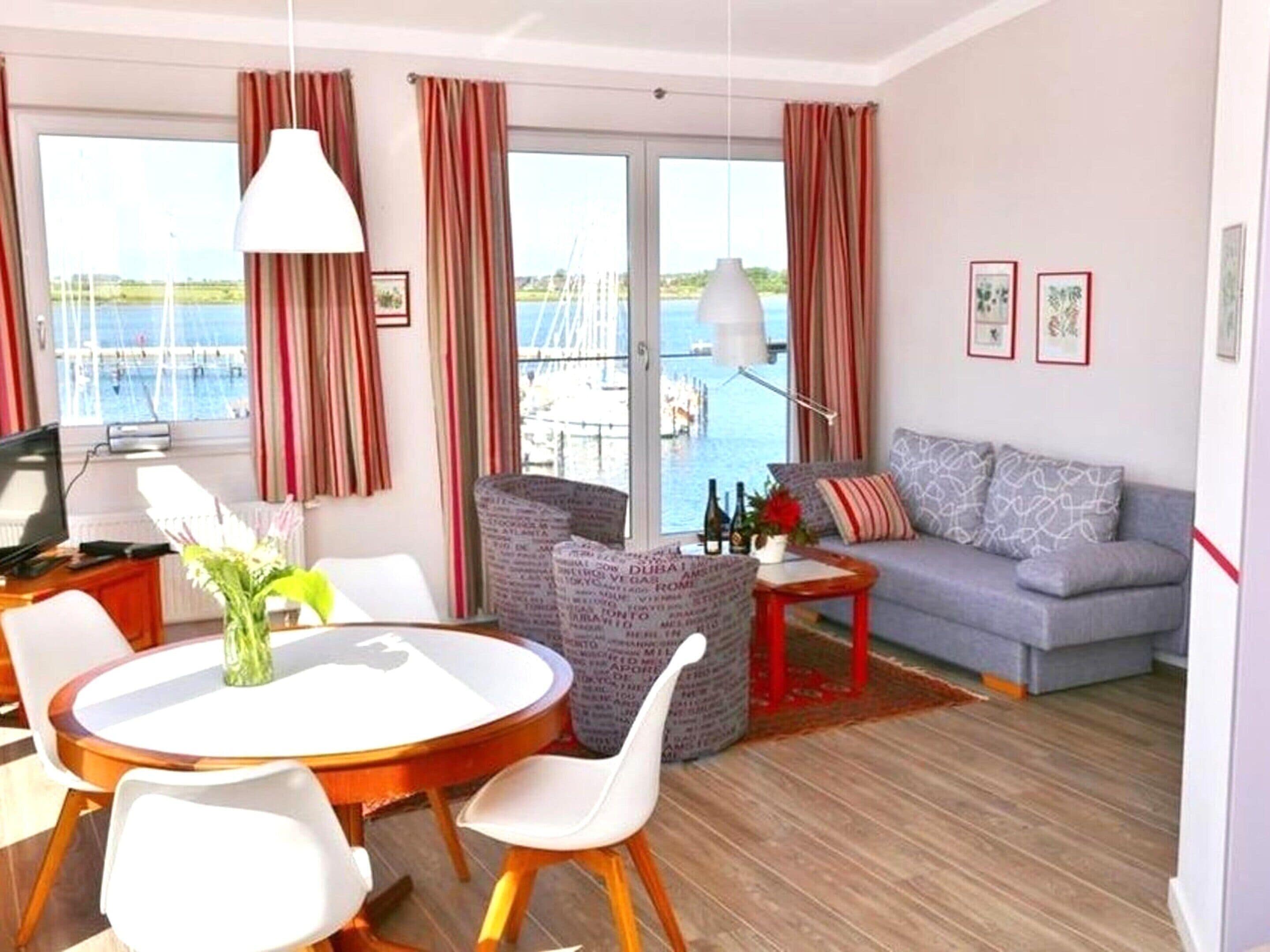 A modern living room with a sofa, a dining table with chairs, a TV, and a view of the sea through large windows.