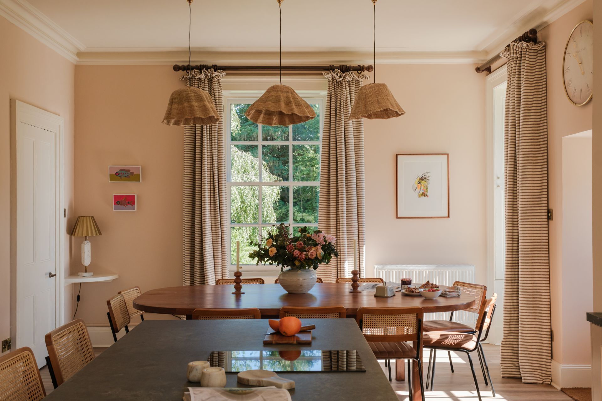 A dining area with a round table, chairs, hanging lamps, a window with curtains, and a clock on the wall.