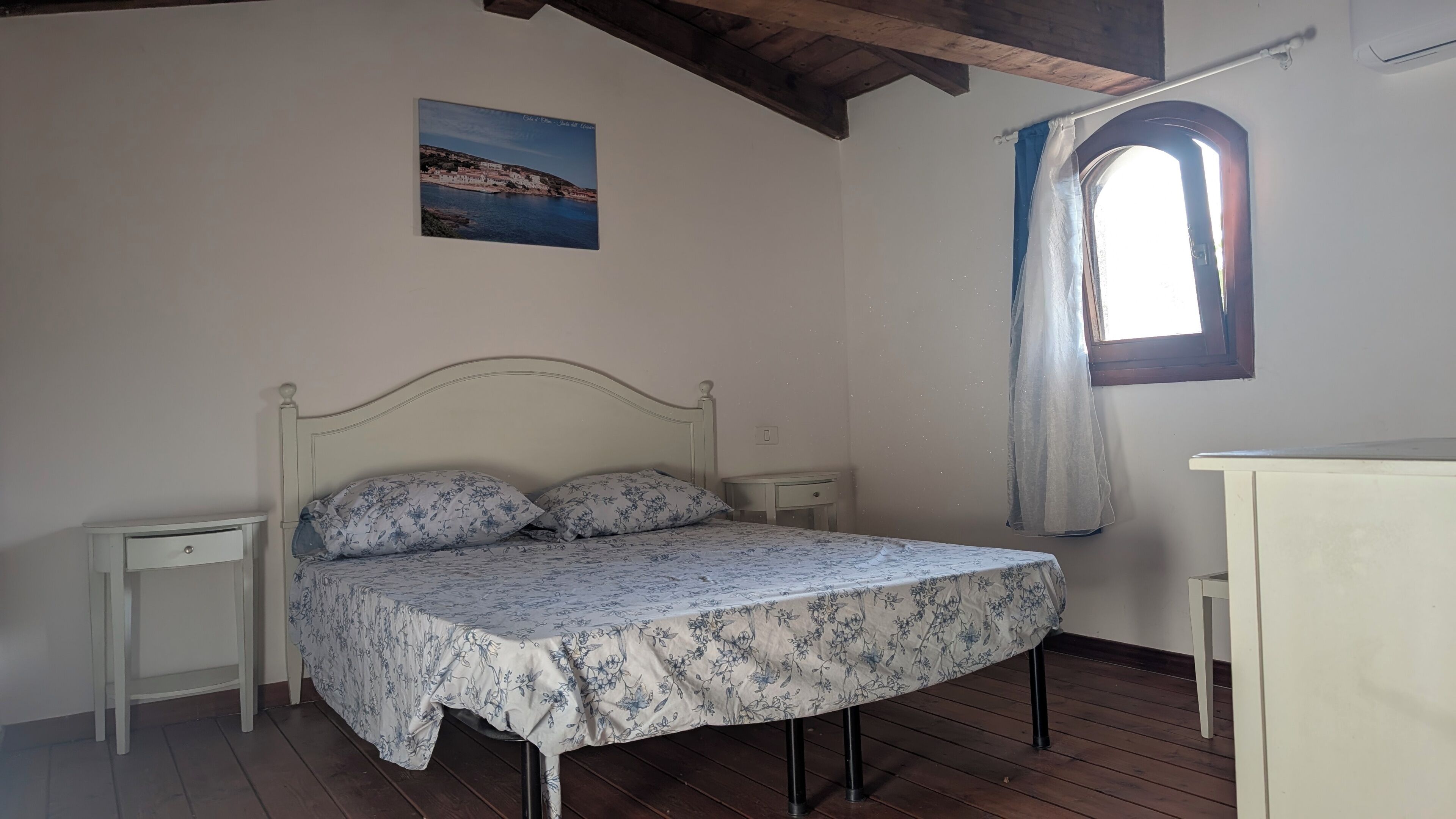 Een slaapkamer met een enkel bed, een nachtkastje, een kleine tafel, een raam met gordijn en een foto aan de muur.
