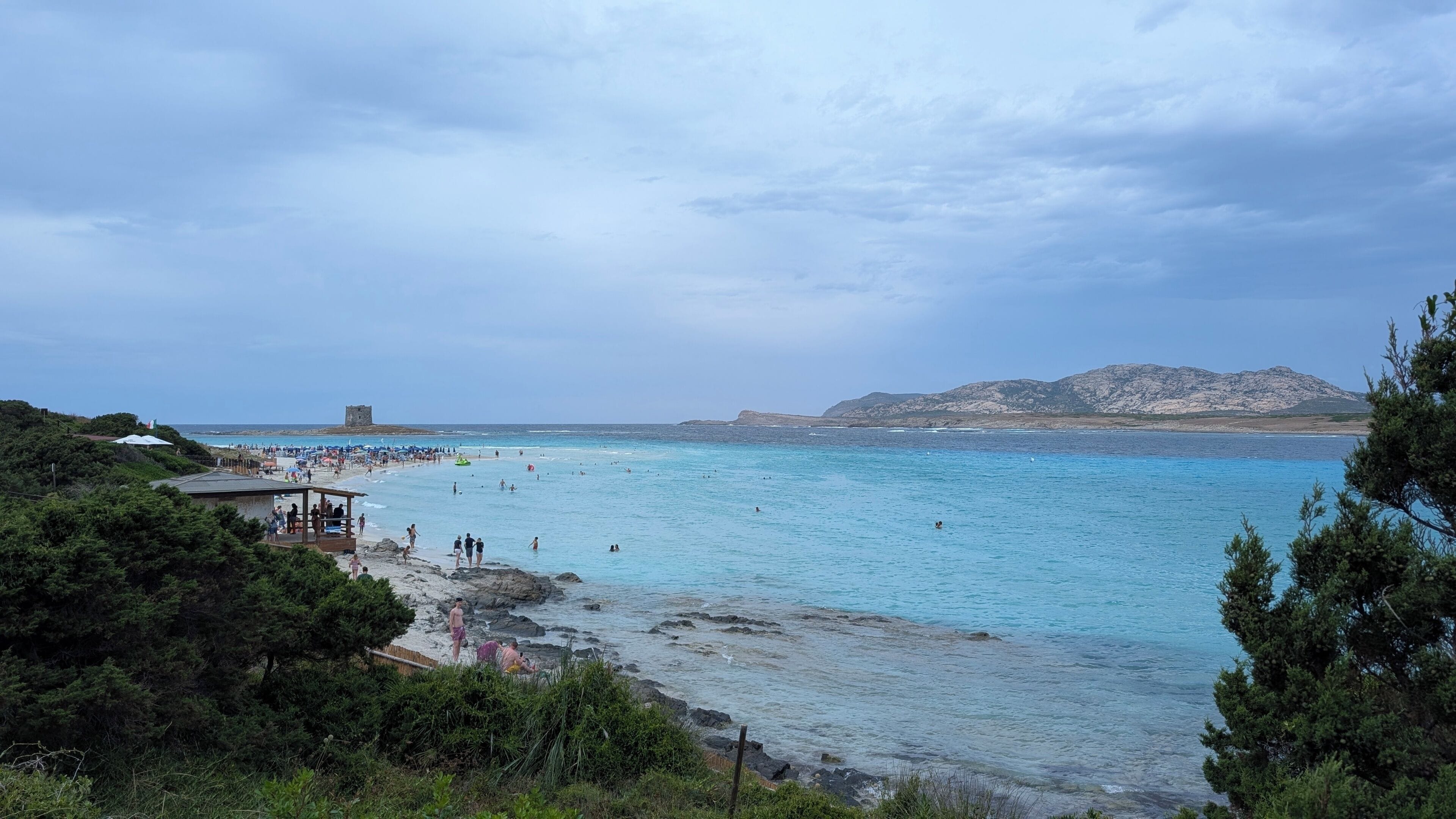 A coastal beach with clear waters, rocky shore, and a distant mountain range.