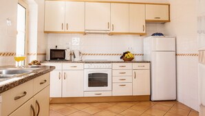 Une cuisine avec des meubles blancs, un four à micro-ondes, un four, un réfrigérateur et un panier de fruits posé sur le plan de travail.