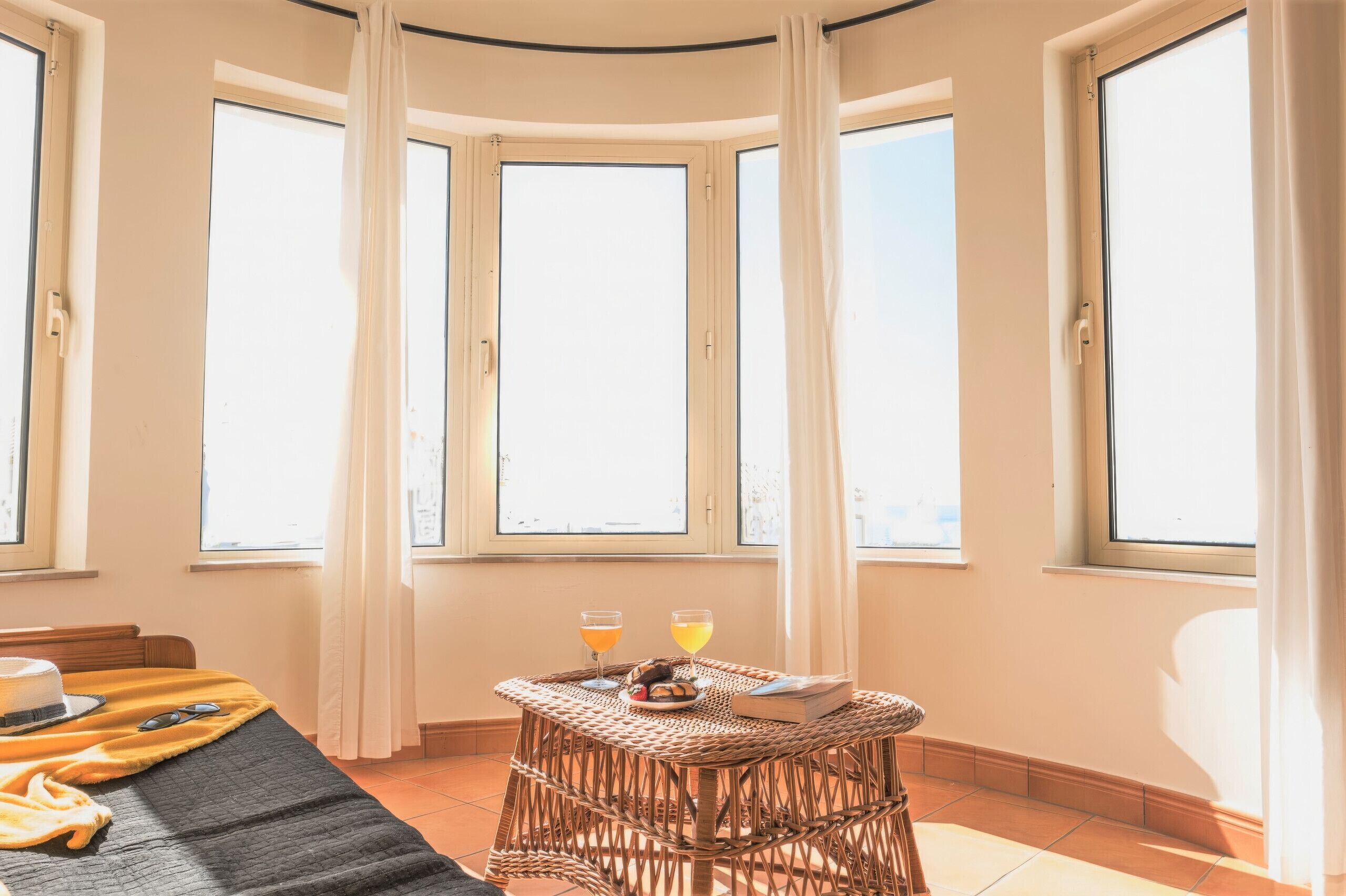 Une pièce avec une baie vitrée en arc de cercle, une table en osier sur laquelle sont posées des boissons et un livre, et un lit avec une couverture et des oreillers.