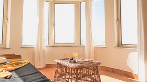 Une pièce avec une baie vitrée en arc de cercle, une table en osier sur laquelle sont posées des boissons et un livre, et un lit avec une couverture et des oreillers.