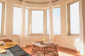 Ein Zimmer mit einem bogenförmigen Erkerfenster, einem Korbtisch mit Getränken und einem Buch sowie einem Bett mit Decke und Kissen.