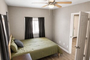 Un dormitori amb llit, ventilador al sostre, finestra amb cortines i porta.
