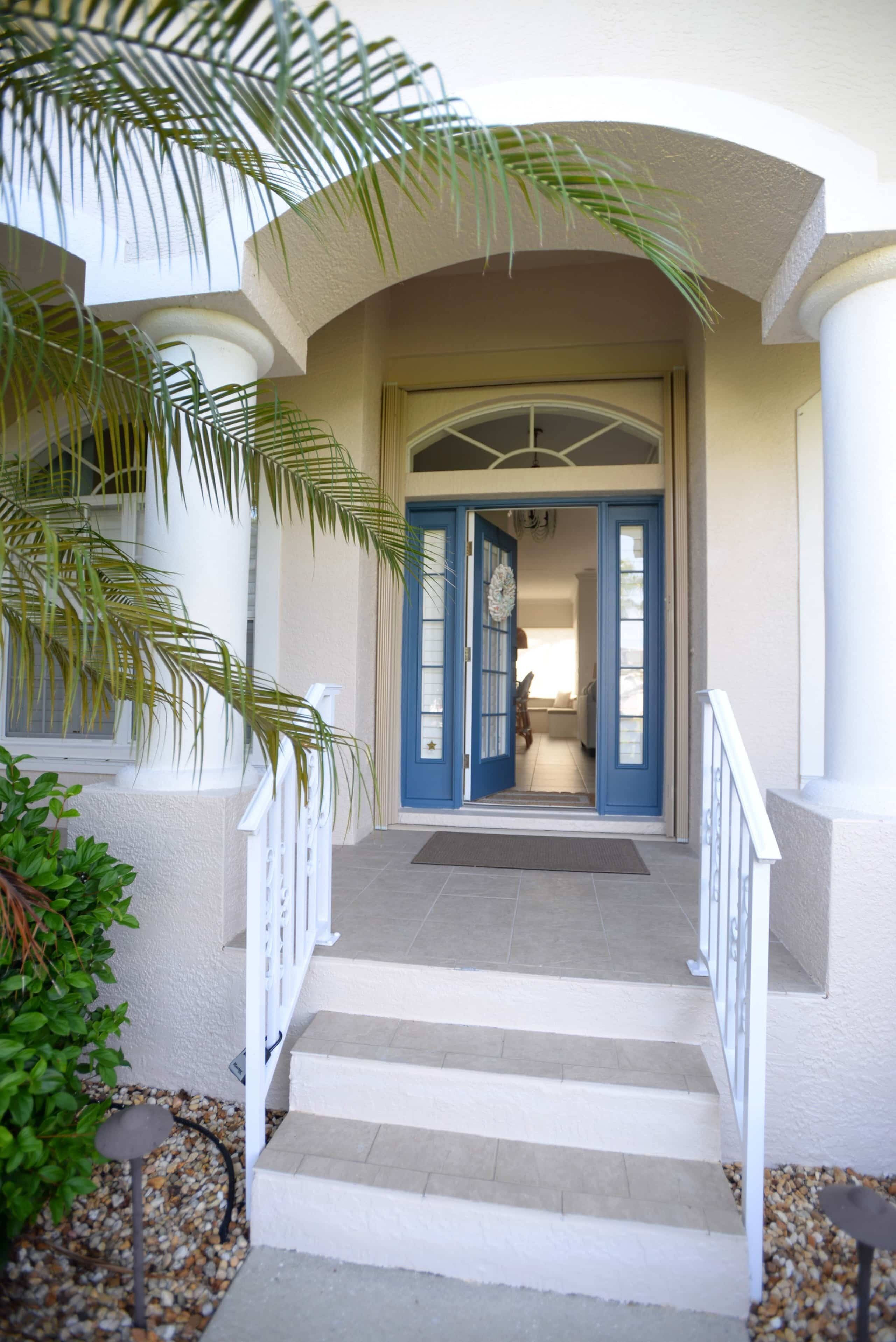 Una escalera conduce a una puerta con marco azul, flanqueada por barandillas blancas y una palmera.