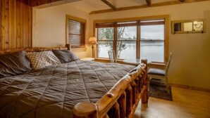 Een kamer met een groot bed, houten hoofdbord, een raam met uitzicht op een wateroppervlak en een klein tafeltje met stoel.
