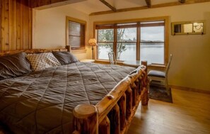 Ein Zimmer mit einem großen Bett, einem Holz-Kopfteil, einem Fenster mit Blick auf ein Gewässer und einem kleinen Tisch mit Stuhl.