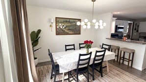 A dining area with a table, chairs, a vase with flowers, a painting on the wall, and a kitchen in the background.
