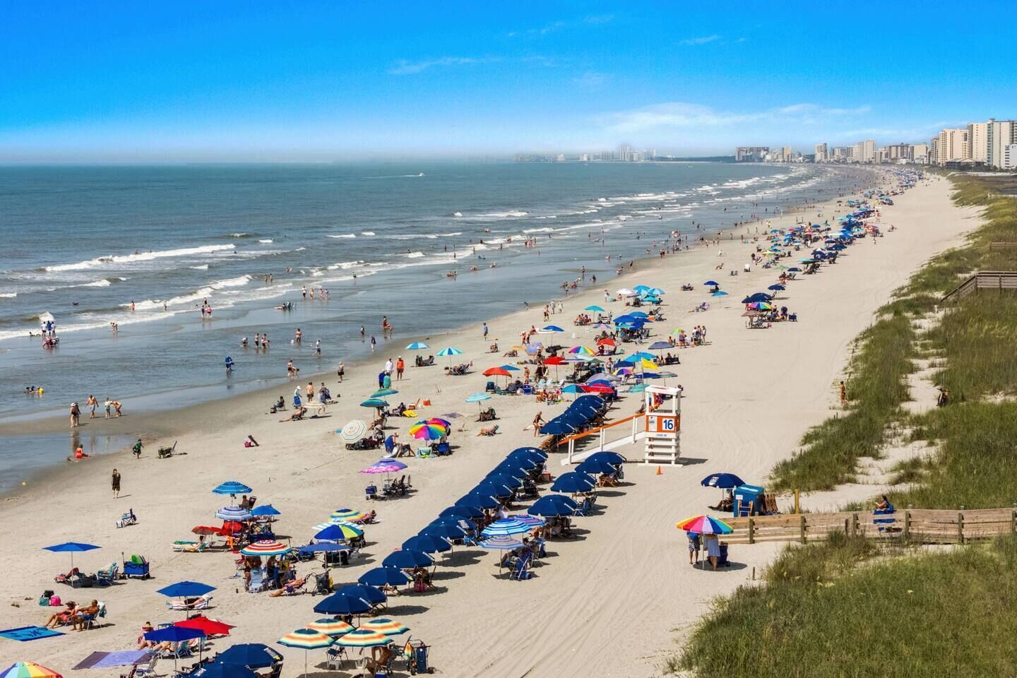 Una playa con hileras de sombrillas y sillas, una torre de salvavidas y un horizonte urbano al fondo.